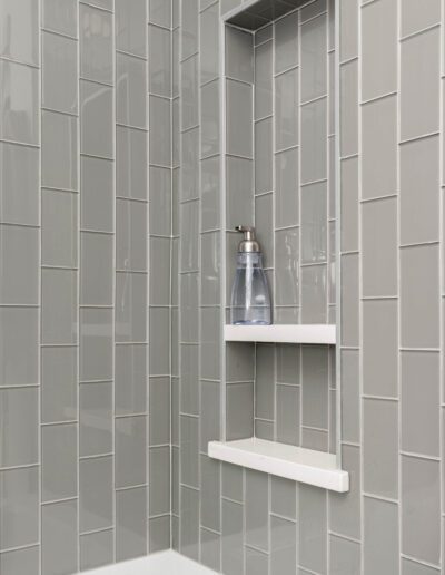 A modern bathroom corner with gray tiles and a built-in shelf holding a single shampoo bottle.