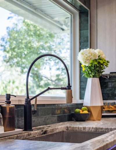 Modern kitchen sink with a high-arc faucet, overlooking a window with a view of greenery, complemented by a vase of white flowers and a bowl of citrus fruits on the countertop.