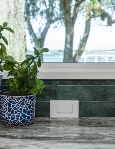 Kitchen counter with a green plant in a white pot, a blue patterned pot with herbs, and a wooden cutting board, overlooking a window with a view of trees.