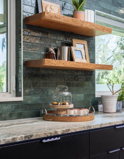 A modern kitchen corner featuring wooden shelves with plants and decor, a countertop with a glass jar of cookies, and a dark tile backsplash.