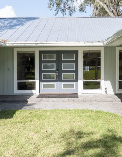 A front view of a house with a gray roof and light blue siding, featuring a double glass door entrance and a small landscaped garden.