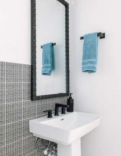 A modern bathroom featuring a white pedestal sink with a black faucet, a decorated mirror, and towels hanging on a wall-mounted rack.