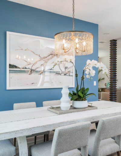 A modern dining room with a white table and chairs, blue accent wall, decorative chandelier, and a beach-themed artwork.