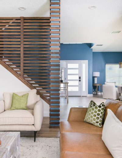 Modern living room with brown leather couch, two white armchairs, and wooden staircase.