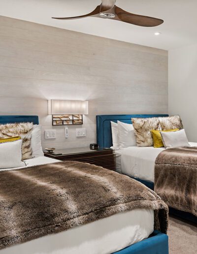 Modern bedroom with two twin beds featuring blue headboards, faux fur blankets, and a ceiling fan.