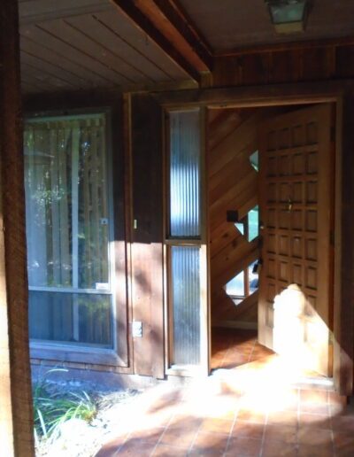 A wooden house entrance with an open door casting a shadow on the floor, surrounded by trees.