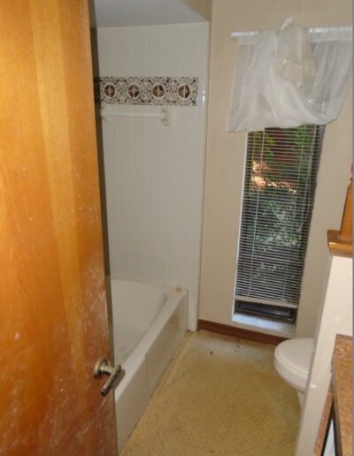 A small bathroom with a white bathtub, toilet, patterned border tiles, and a window with blinds partially closed.